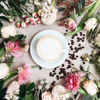 卡布奇诺填充白色陶瓷咖啡杯在白色陶瓷茶托上，周围有各种各样的花和咖啡豆